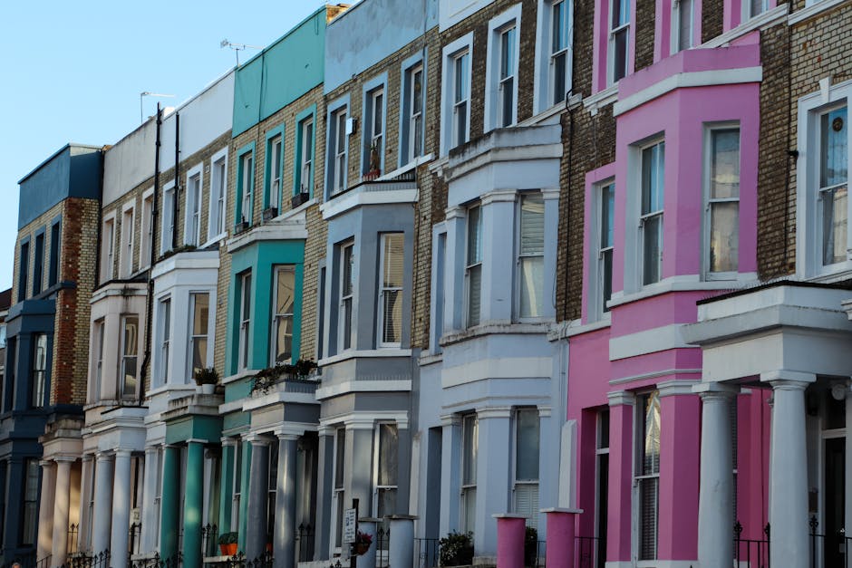 A row of Victorian-style terraced houses with colourful facades, including shades of pink, blue, and teal, situated on a residential street in Clapham. The buildings feature bay windows, white window frames, and decorative cornices. Some houses have small front gardens with planters and wrought iron fences, while others are directly accessible from the pavement. The image is taken during daylight with clear weather, showing the height and uniformity of the terraced architecture. This street scene offers a visual context for house relocations or furniture transport, highlighting the exterior of homes that may require professional moving services by companies like Man with Van Clapham, especially during packing and home moving processes.