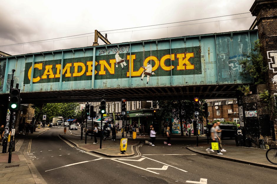 A street scene under a large, weathered railway bridge with a faded greenish-blue paint, displaying the words '20 FOOT CAMDEN LOCK' in bold yellow lettering. The bridge features two hanging figures painted on its facade, one swinging and the other hanging from a rope. Below, pedestrians are walking across a busy intersection, some bundled up in light clothing, with a mixture of modern buildings and graffiti-covered walls visible in the background. Several traffic lights are illuminated green, and the road includes striped bicycle lanes and pedestrian crossings. On the right side, a black brick building is partially visible amidst greenery. The scene captures a typical urban environment in London, relevant to city-based house removals and moving services, with the bridge indicating a popular area for furniture transport and home relocation activities. The presence of pedestrians and street activity reflects the logistics involved in loading and unloading household items in a busy city setting, managed by companies like Man with Van Clapham.